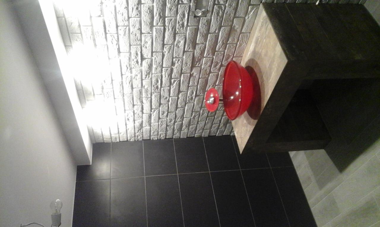 Nowoczesna łazienka z czarnymi płytkami podłogowymi, ścianą z białej cegły dekoracyjnej i umywalką w intensywnym czerwonym kolorze na drewnianym blacie.