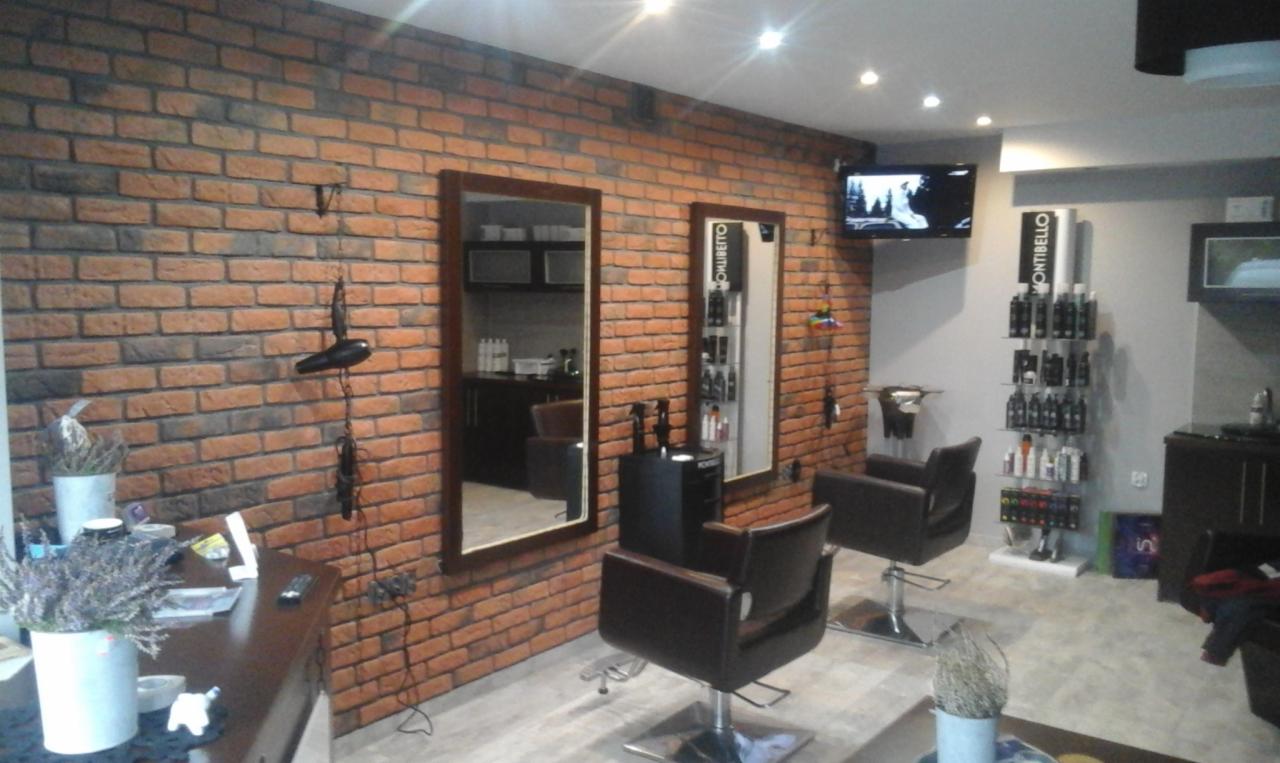 Salon fryzjerski z ceglaną ścianą, dwoma lustrami w drewnianych ramach, stanowiskami fryzjerskimi z brązowymi fotelami, półką z kosmetykami Montibello i telewizorem na ścianie.