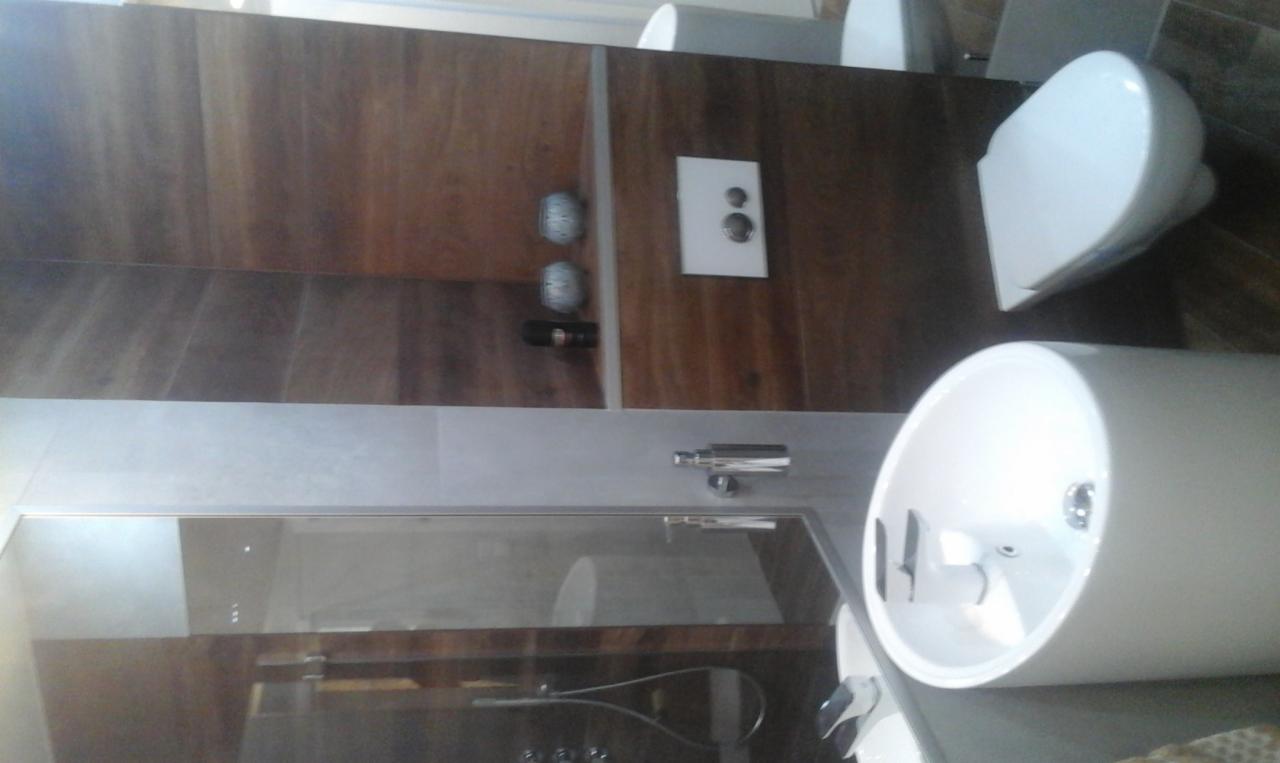 Nowoczesna łazienka z drewnianymi panelami ściennymi, białą umywalką nablatową, wiszącą toaletą i szklaną kabiną prysznicową.