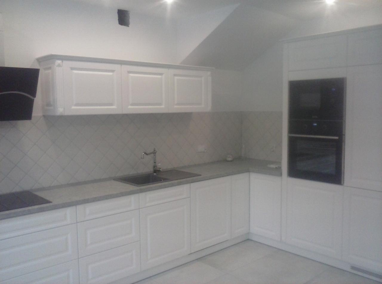 Jasna kuchnia z białymi, frezowanymi szafkami, szarym blatem i płytkami ułożonymi w romby, widziana pod kątem w słabym oświetleniu.