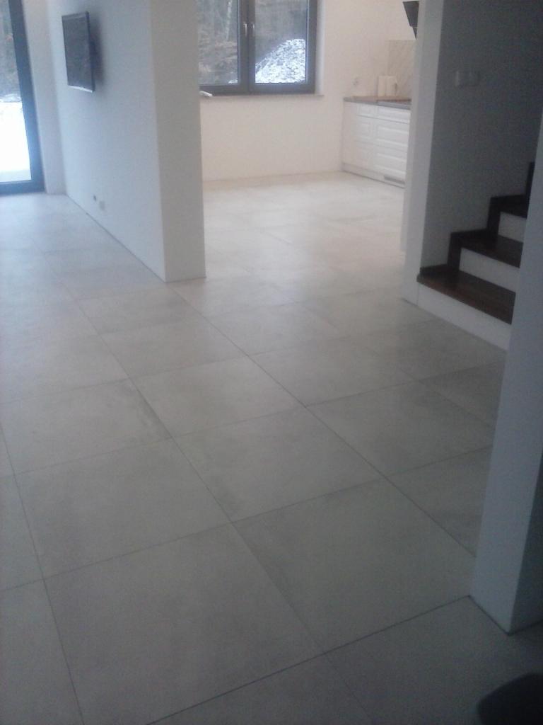 Jasnoszara, matowa podłoga z płytek w nowym, pustym wnętrzu. Widoczny fragment schodów z drewnianymi stopniami, białe ściany i okno z widokiem na zaśnieżony krajobraz.