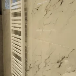 Biały, drabinkowy grzejnik łazienkowy zamontowany na ścianie wyłożonej jasnymi, marmurkowymi płytkami w nowoczesnej łazience.