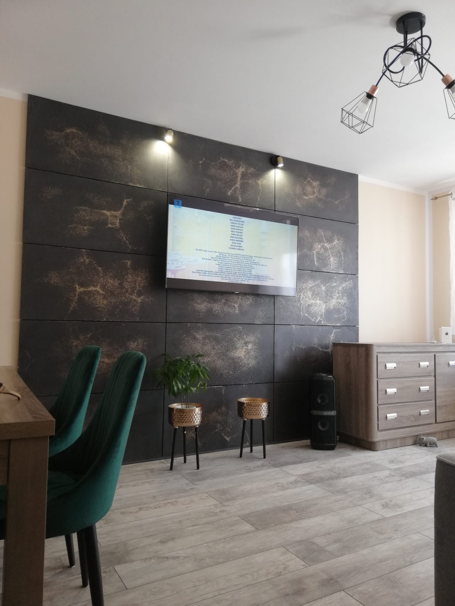 Ściana telewizyjna wykończona panelami imitującymi beton z delikatnym złotym wzorem, podświetlona punktowymi lampami, z widocznymi elementami wystroju wnętrza: zielone krzesła, komoda i głośnik.