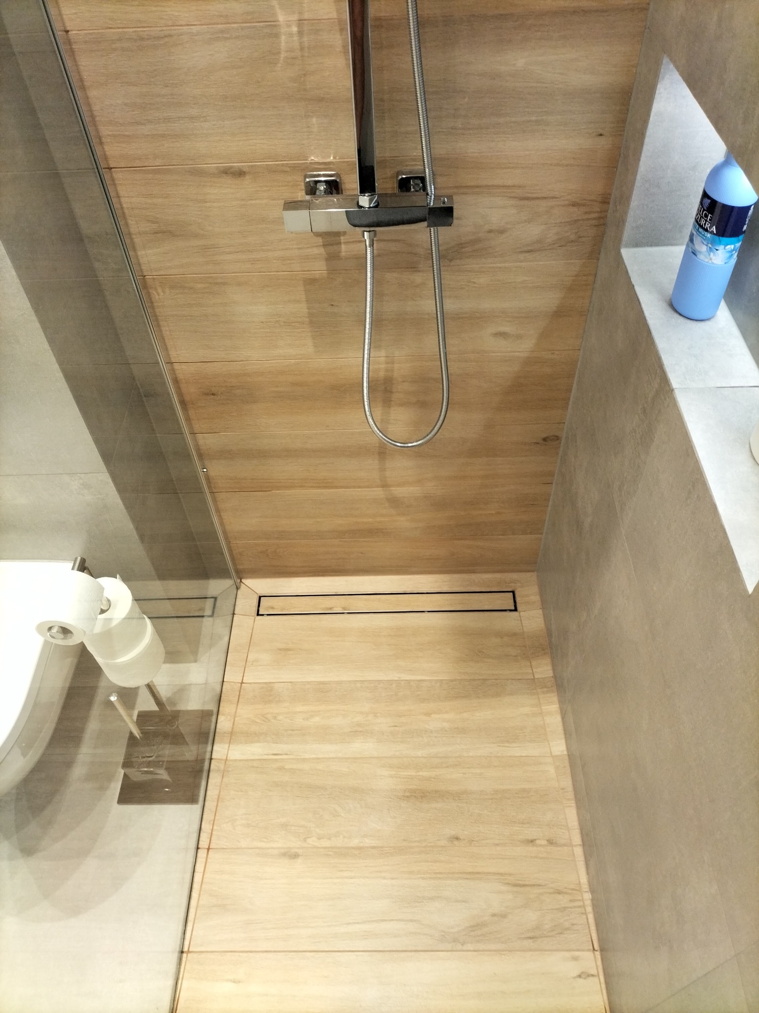 Nowoczesna kabina prysznicowa z drewnopodobnymi płytkami na ścianach i podłodze, odpływ liniowy, minimalistyczna armatura, półka z błękitnym płynem do kąpieli.