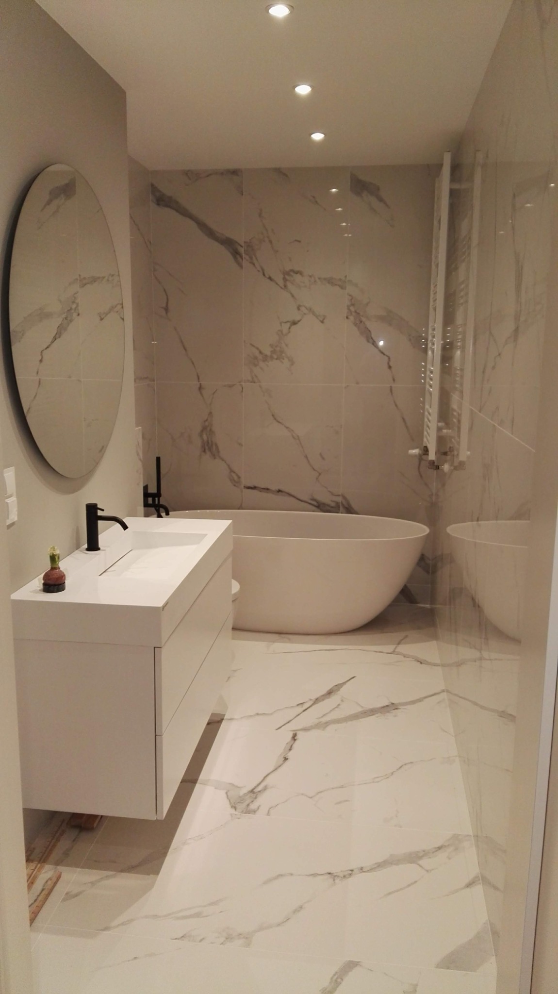 Nowoczesna łazienka z wanną wolnostojącą, umywalką nablatową z czarną armaturą i okrągłym lustrem, wykończona płytkami imitującymi biały marmur z szarymi żyłami.