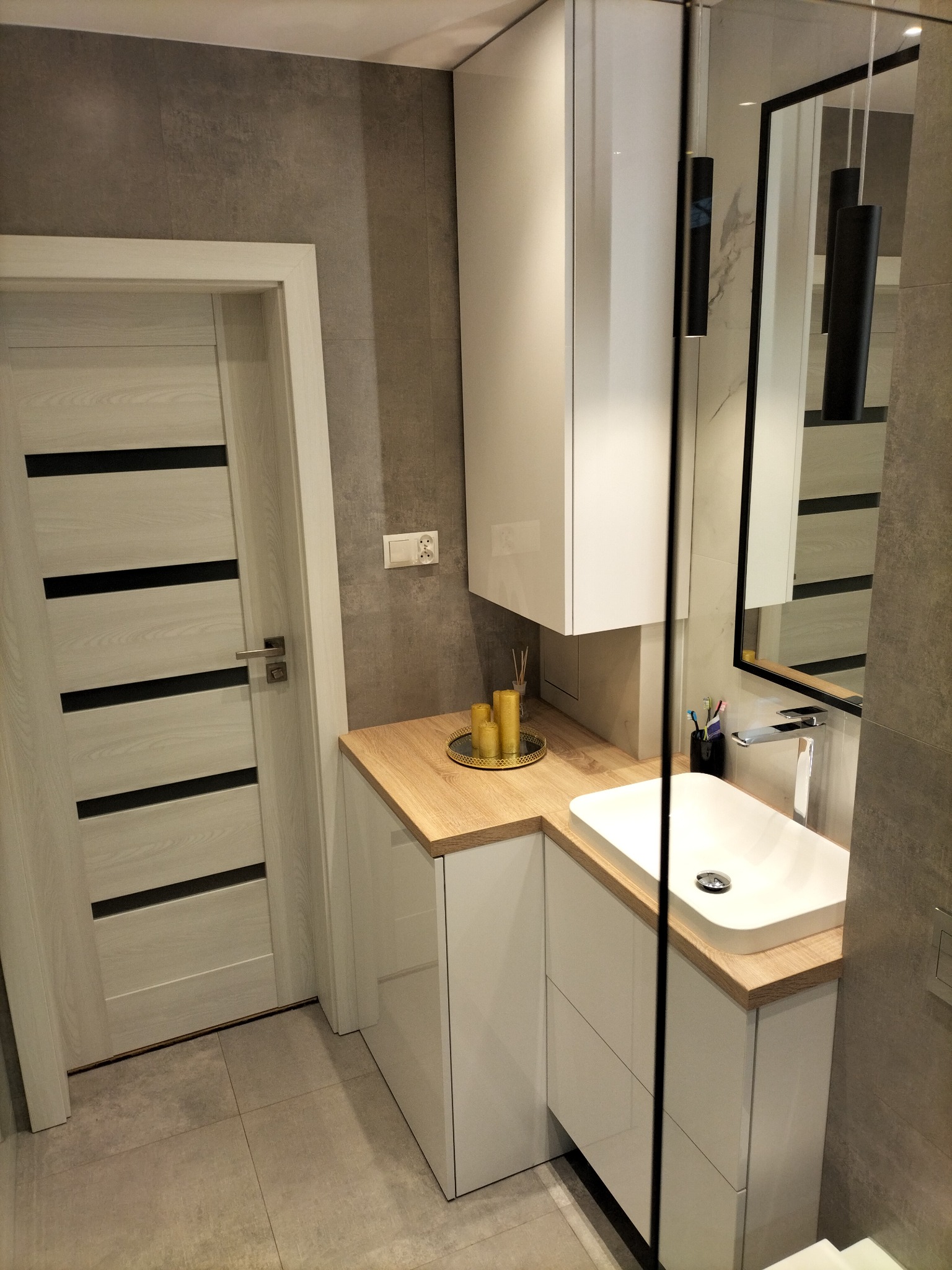 Nowoczesna łazienka z umywalką nablatową, szafką z białymi frontami i blatem w kolorze drewna, szara ściana z płytek, drzwi z poziomymi czarnymi paskami i wisząca lampa w czarnej tubie.