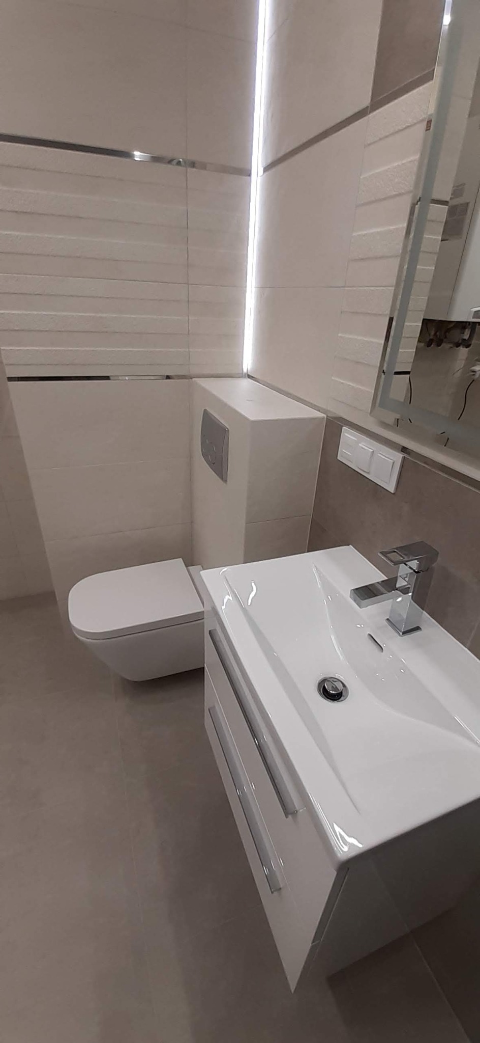 Nowoczesna łazienka z wiszącą toaletą i białą szafką z umywalką, chromowana bateria, płytki imitujące kamień z poziomymi paskami, oświetlenie LED w pionowej szczelinie.