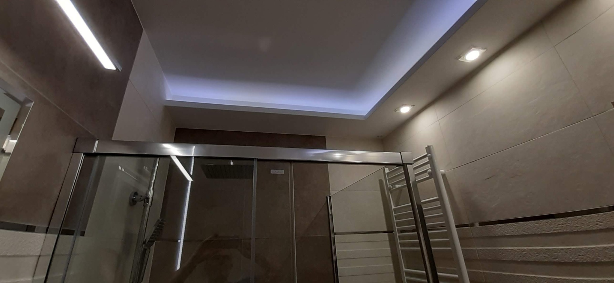 Nowoczesna łazienka z kabiną prysznicową z przesuwnymi drzwiami, podwieszanym sufitem z oświetleniem LED i grzejnikiem drabinkowym.