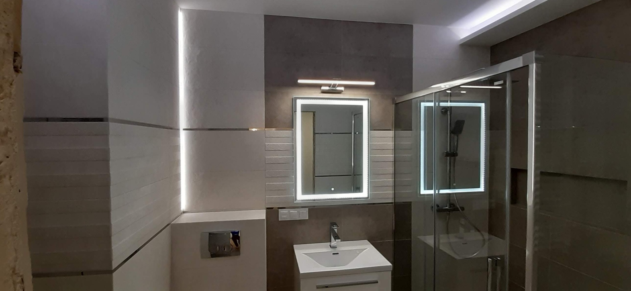 Nowoczesna łazienka z podświetlanym lustrem, kabiną prysznicową ze szklanymi drzwiami i umywalką z szafką, wykończona płytkami w odcieniach bieli i szarości z poziomymi paskami dekoracyjnymi.