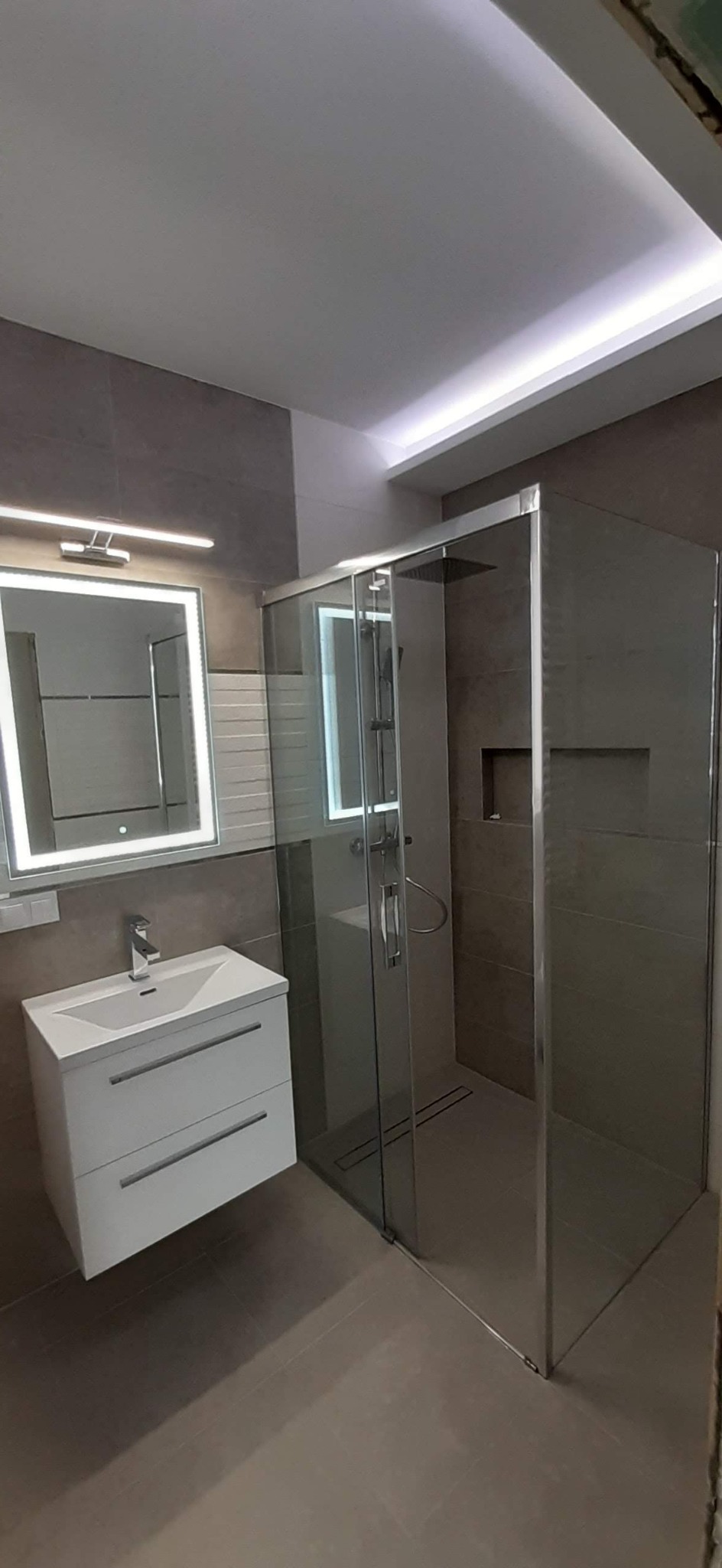 Nowoczesna łazienka z kabiną prysznicową typu walk-in z szarymi płytkami, umywalką z szafką i podświetlanym lustrem, oświetlenie sufitowe LED.