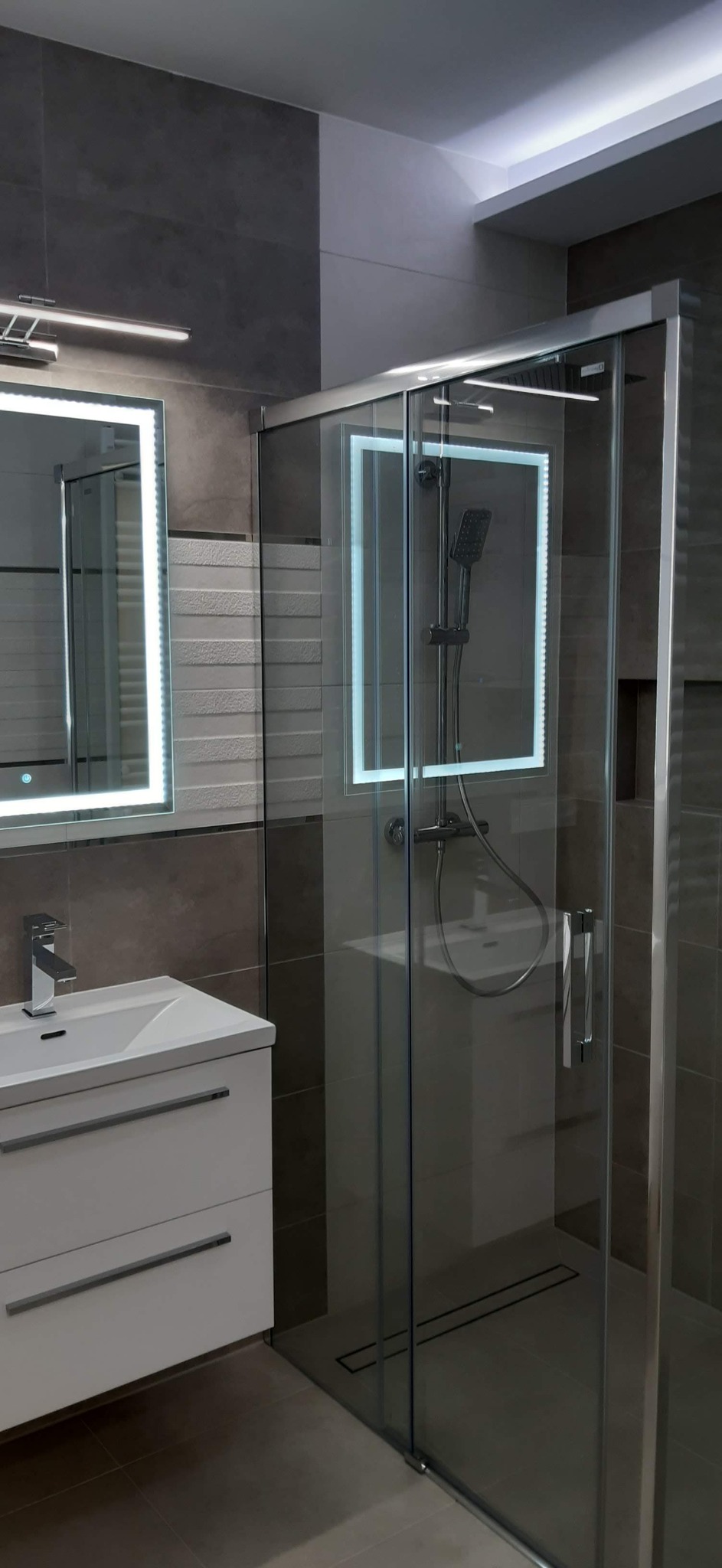 Nowoczesna łazienka z kabiną prysznicową ze szklanymi drzwiami przesuwnymi, umywalką z szafką w kolorze białym i podświetlanymi lustrami w kształcie prostokąta, wykończona płytkami w odcieniach...