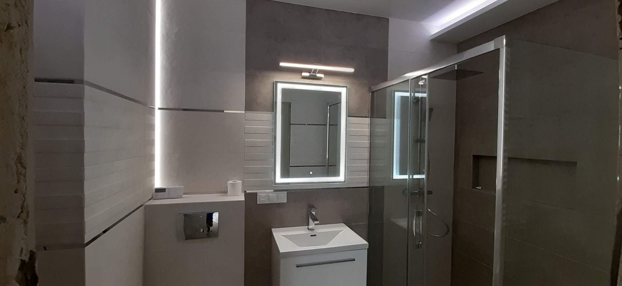 Nowoczesna łazienka z podświetlanym lustrem, kabiną prysznicową z szarymi płytkami i ukrytą spłuczką toaletową, wykończona w odcieniach bieli i szarości.