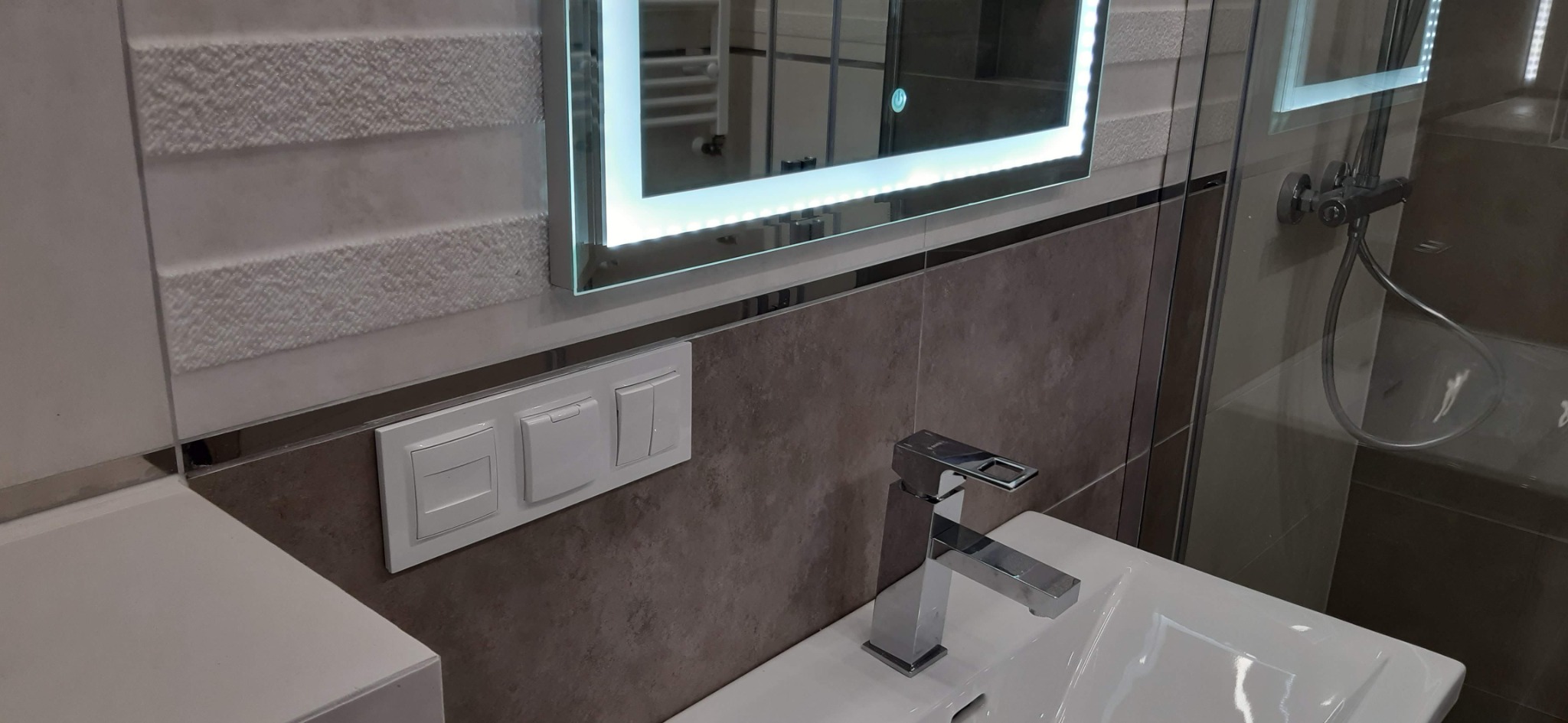 Nowoczesna łazienka z umywalką, baterią, podświetlanym lustrem i przełącznikami na ścianie wykończonej płytkami w dwóch odcieniach.