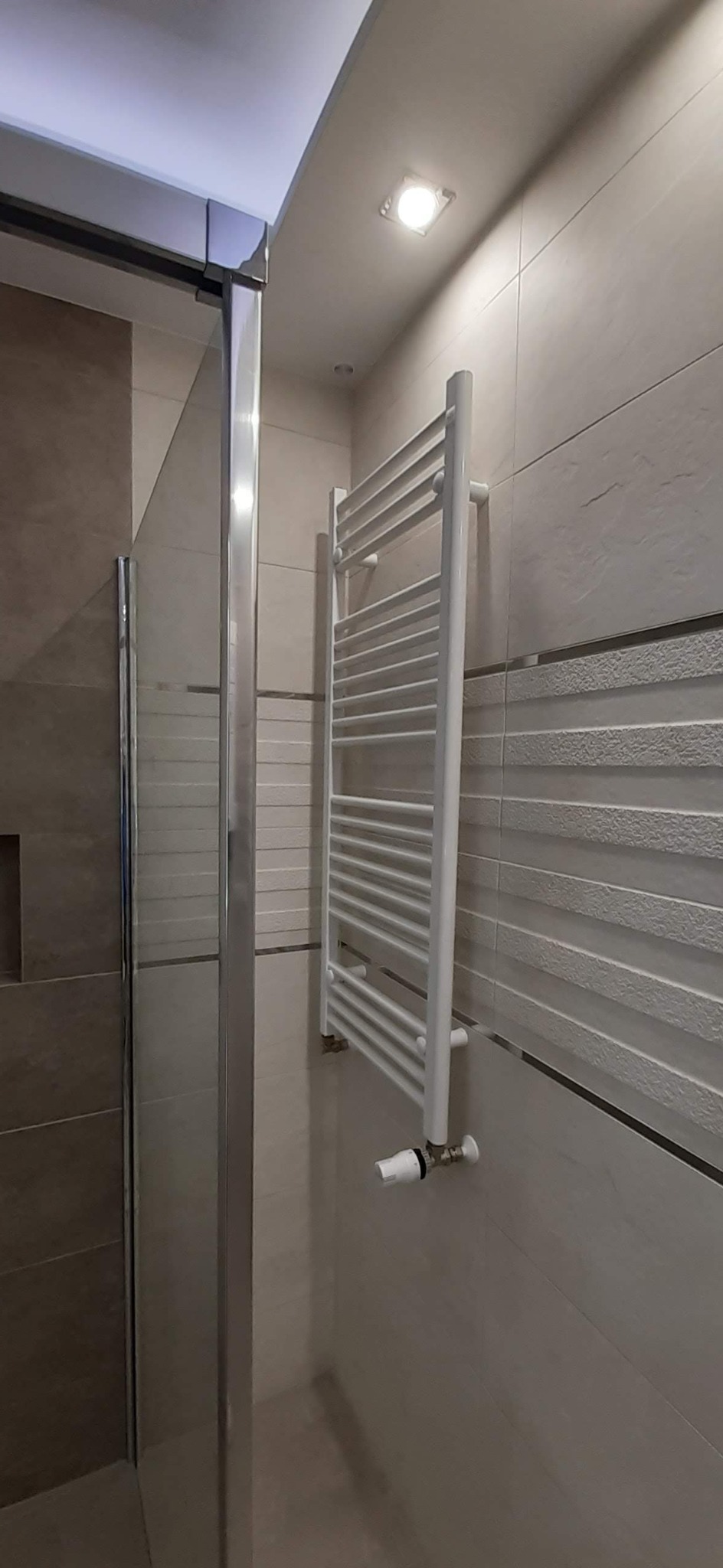 Biały grzejnik drabinkowy zamontowany na beżowej ścianie z poziomymi żłobieniami, widoczny fragment kabiny prysznicowej z chromowanymi profilami.