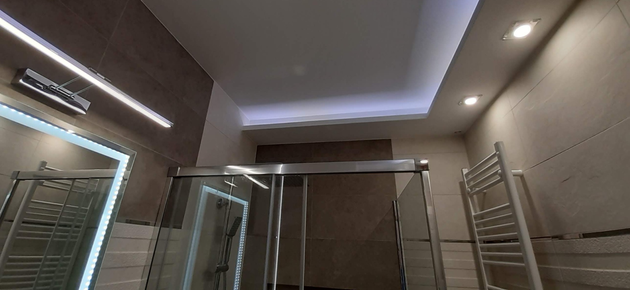 Nowoczesna łazienka z kabiną prysznicową, oświetleniem LED w lustrze i podwieszanym suficie, oraz grzejnikiem drabinkowym na ścianie wyłożonej płytkami w odcieniach beżu i brązu.