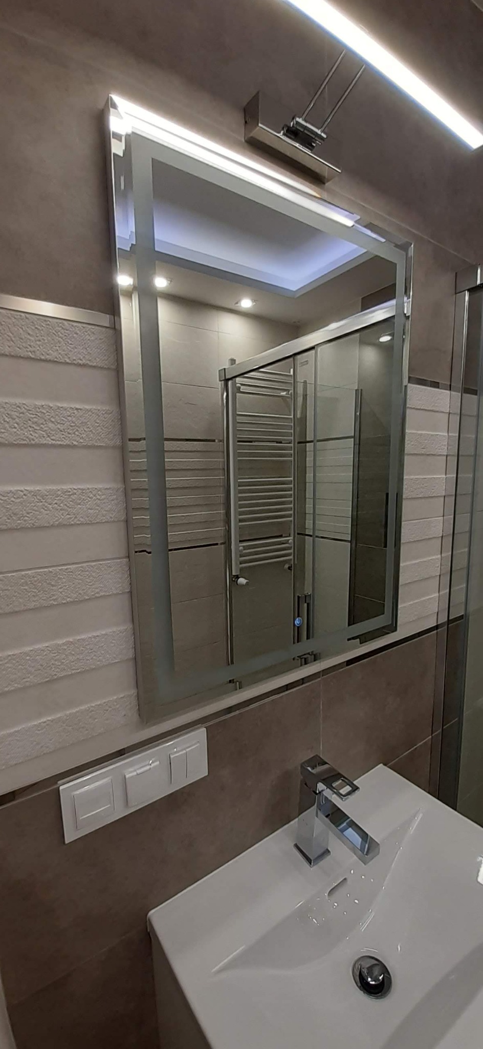 Nowoczesna łazienka z podświetlanym lustrem, umywalką z chromowaną baterią i dekoracyjną ścianą z fakturą, odzwierciedlająca kabinę prysznicową z grzejnikiem drabinkowym.