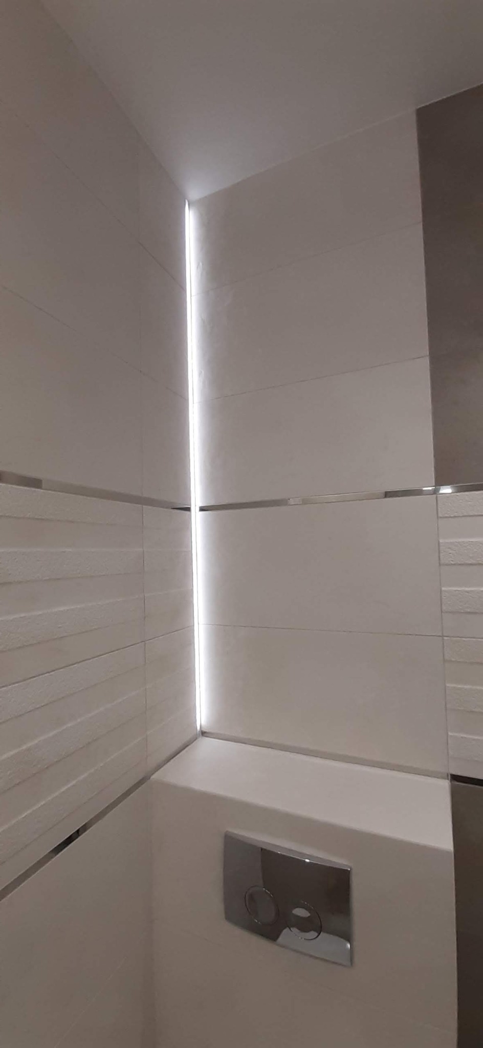 Nowoczesna łazienka z białymi, strukturalnymi płytkami, oświetleniem LED w pionowej szczelinie i chromowanym przyciskiem spłuczki.