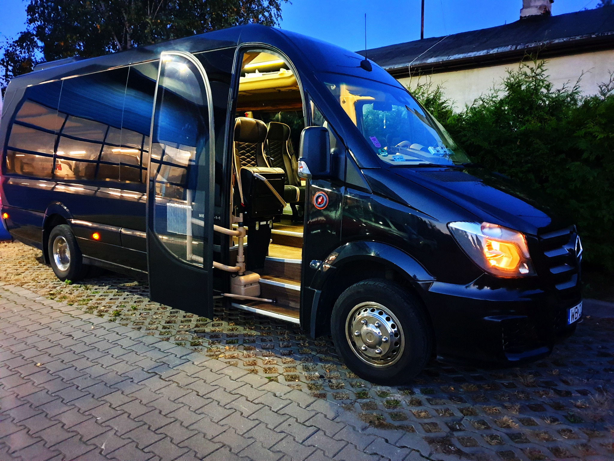 Czarny, luksusowy bus z otwartymi drzwiami wejściowymi, widoczne skórzane fotele pasażerskie wewnątrz, zaparkowany na betonowej kostce.