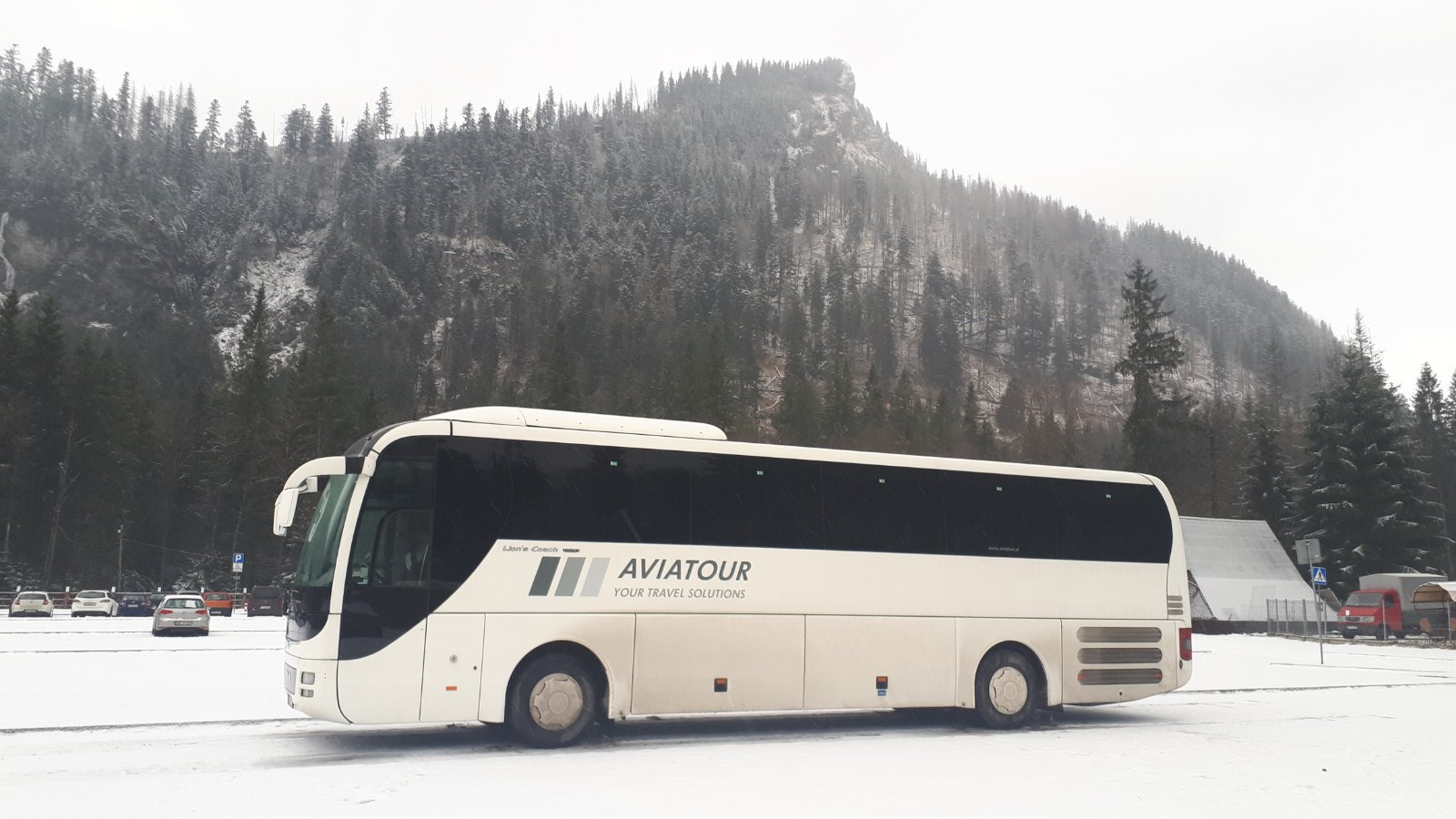 Biały autokar Aviatour na śnieżnym parkingu z zalesioną górą w tle, widoczne zaparkowane samochody i budynki.