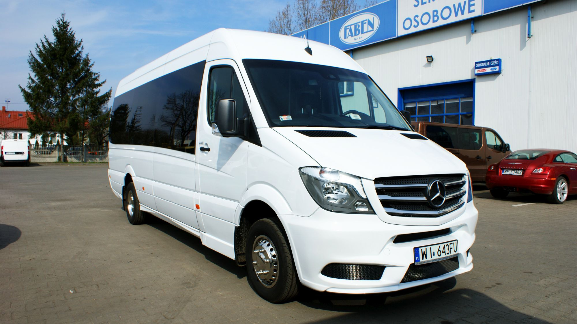 Biały, lśniący bus Mercedes Sprinter z ciemnymi szybami, zaparkowany przed budynkiem serwisu samochodowego z logo 'Faben Serwis Osobowe', widoczne inne pojazdy w tle.