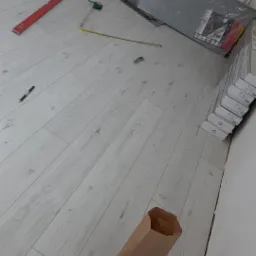 Montaż paneli podłogowych: widoczny poziomica, miarka zwijana, nożyk i paczki paneli, brązowa torba papierowa na podłodze.