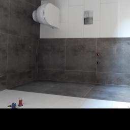 Usługi remontowo-wykonczeniowe MB-BUD - Wnętrze łazienki w trakcie remontu z widoczną białą podwieszaną toaletą, szarą glazurą ścienną, białymi płytkami podłogowymi i wystającymi rurami instalacji wodnej zabezpieczonymi czerwonymi...