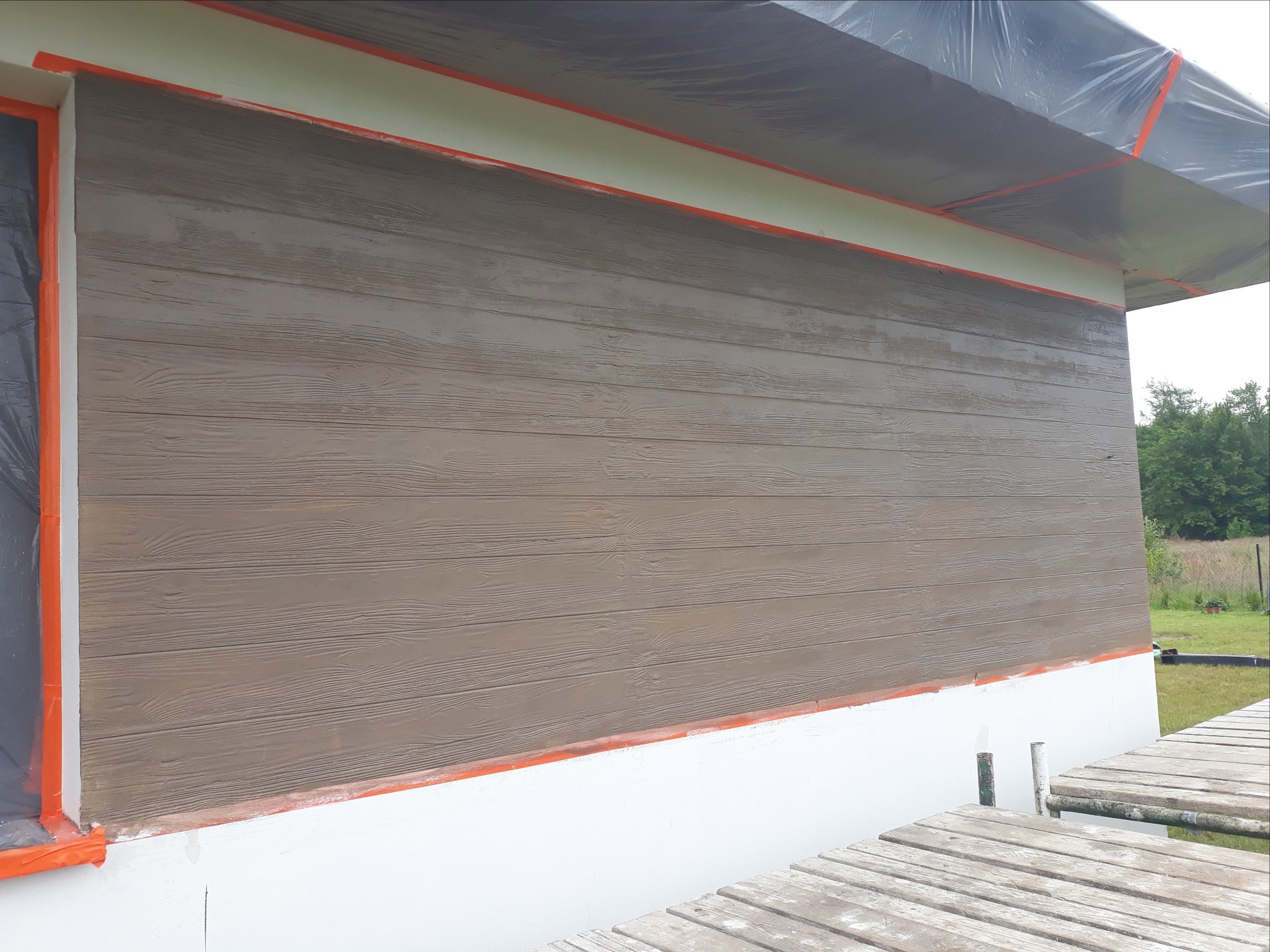 Fragment elewacji budynku z brązową okładziną imitującą drewno, zabezpieczone okno folią i taśmą malarską, widoczne rusztowanie.