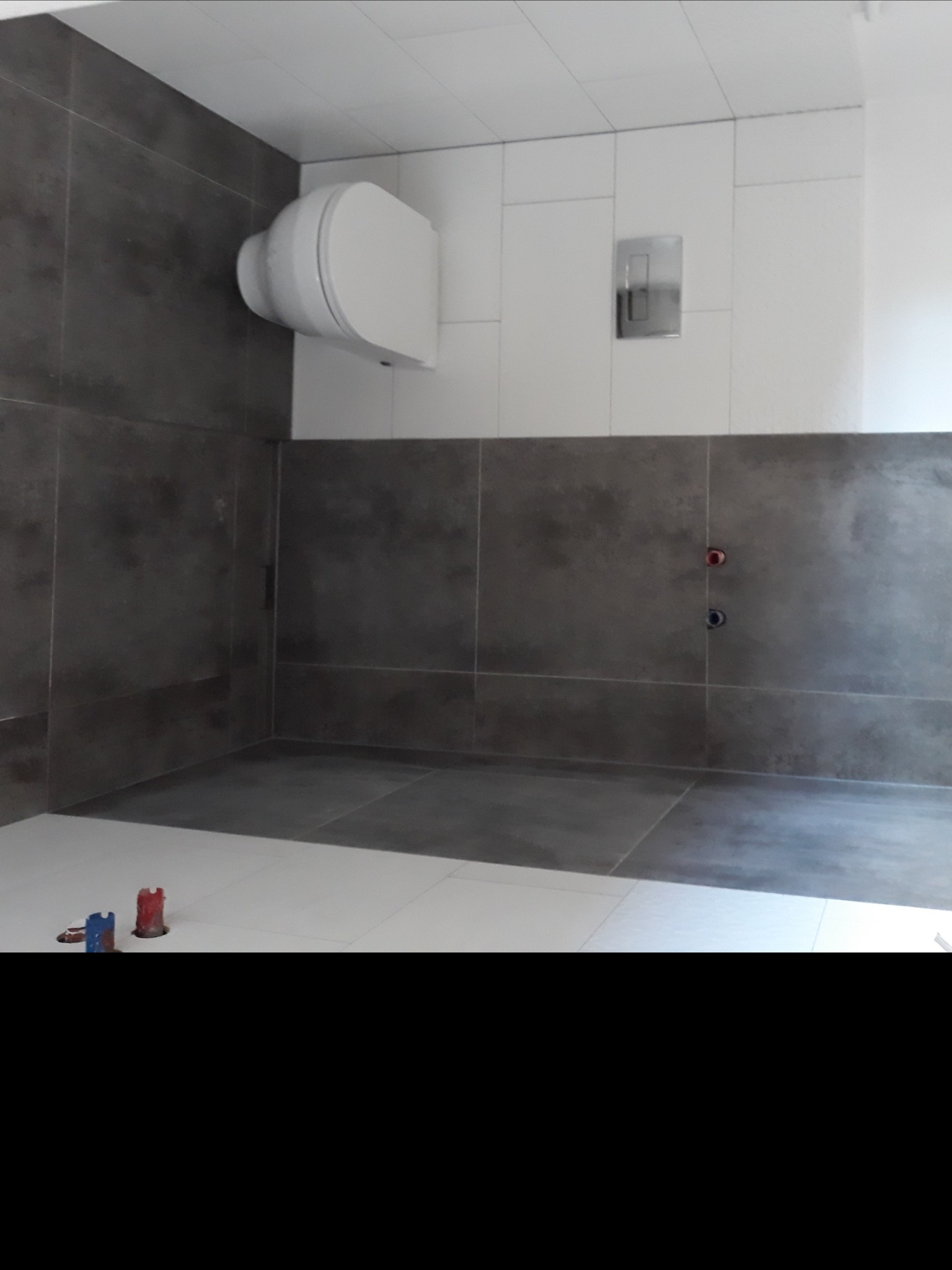 Wnętrze łazienki w trakcie remontu z widoczną białą podwieszaną toaletą, szarą glazurą ścienną, białymi płytkami podłogowymi i wystającymi rurami instalacji wodnej zabezpieczonymi czerwonymi...