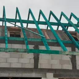 Surowa konstrukcja więźby dachowej z impregnowanego drewna na ścianach z betonu komórkowego, widok z dołu na tle pochmurnego nieba.