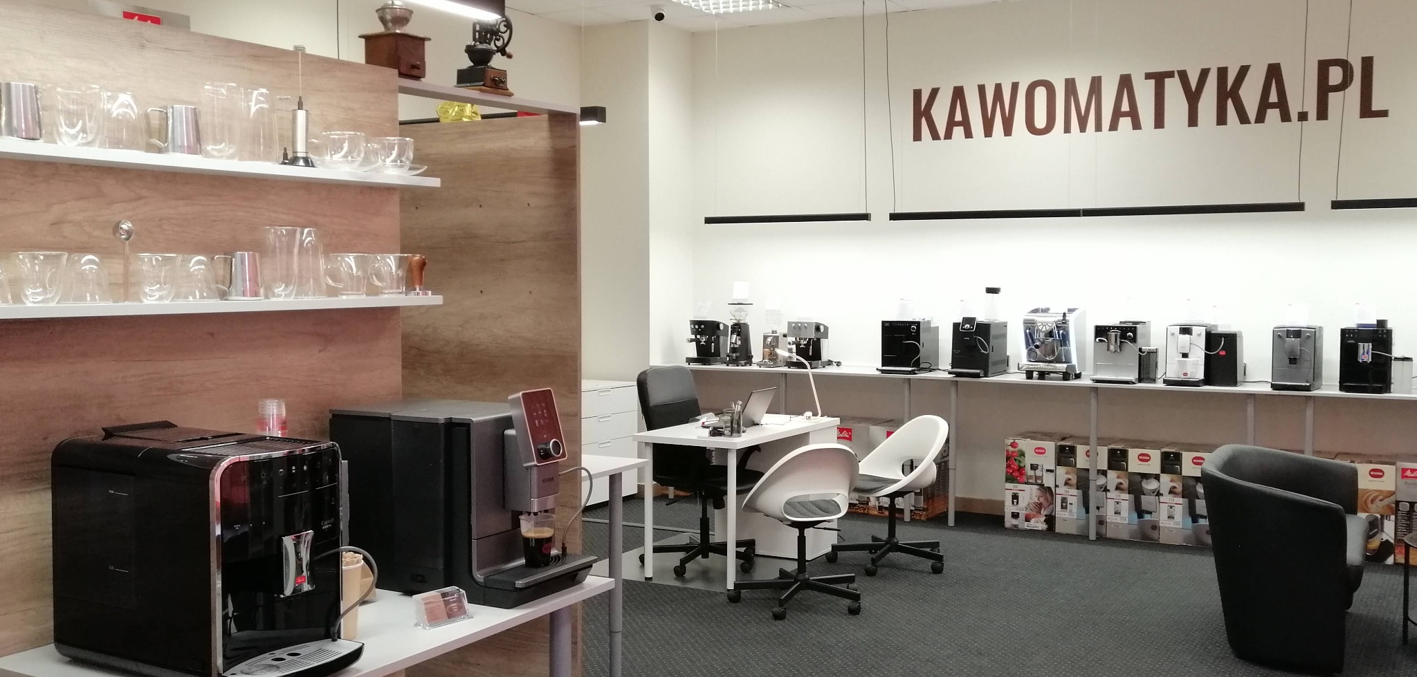 Ekspozycja różnych modeli ekspresów do kawy w salonie sprzedaży, widoczne ekspresy automatyczne, akcesoria do kawy, logo Kawomatyka.pl na ścianie.