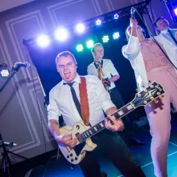 Energiczne ujęcie gitarzysty z wyciągniętym językiem podczas występu na scenie z saksofonistą i wokalistką w tle, oświetlenie sceniczne w kolorach niebieskim i zielonym.