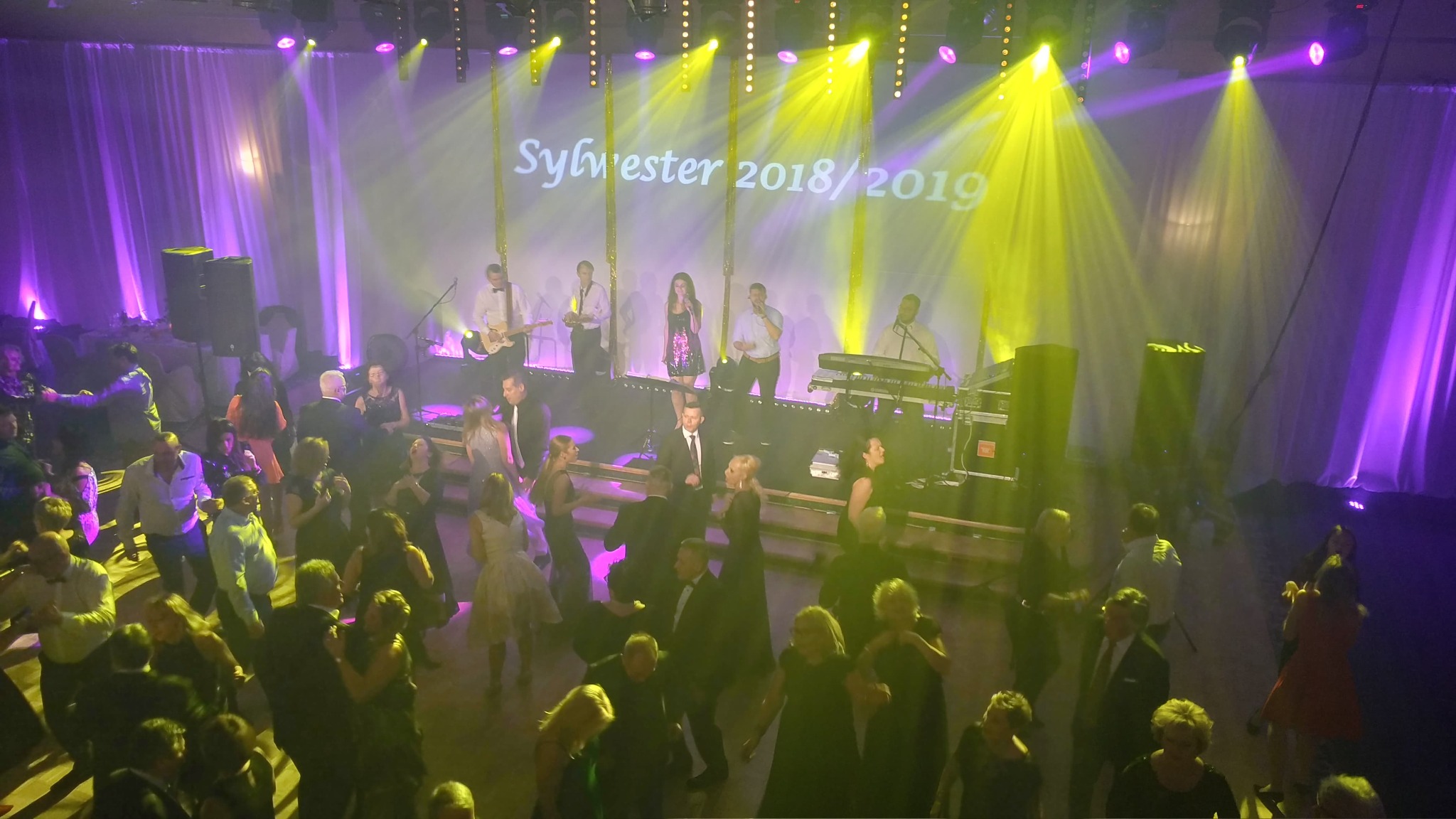 Sylwester 2018/2019: Zespół muzyczny gra na scenie oświetlonej żółtymi i fioletowymi światłami, podczas gdy goście tańczą na parkiecie.