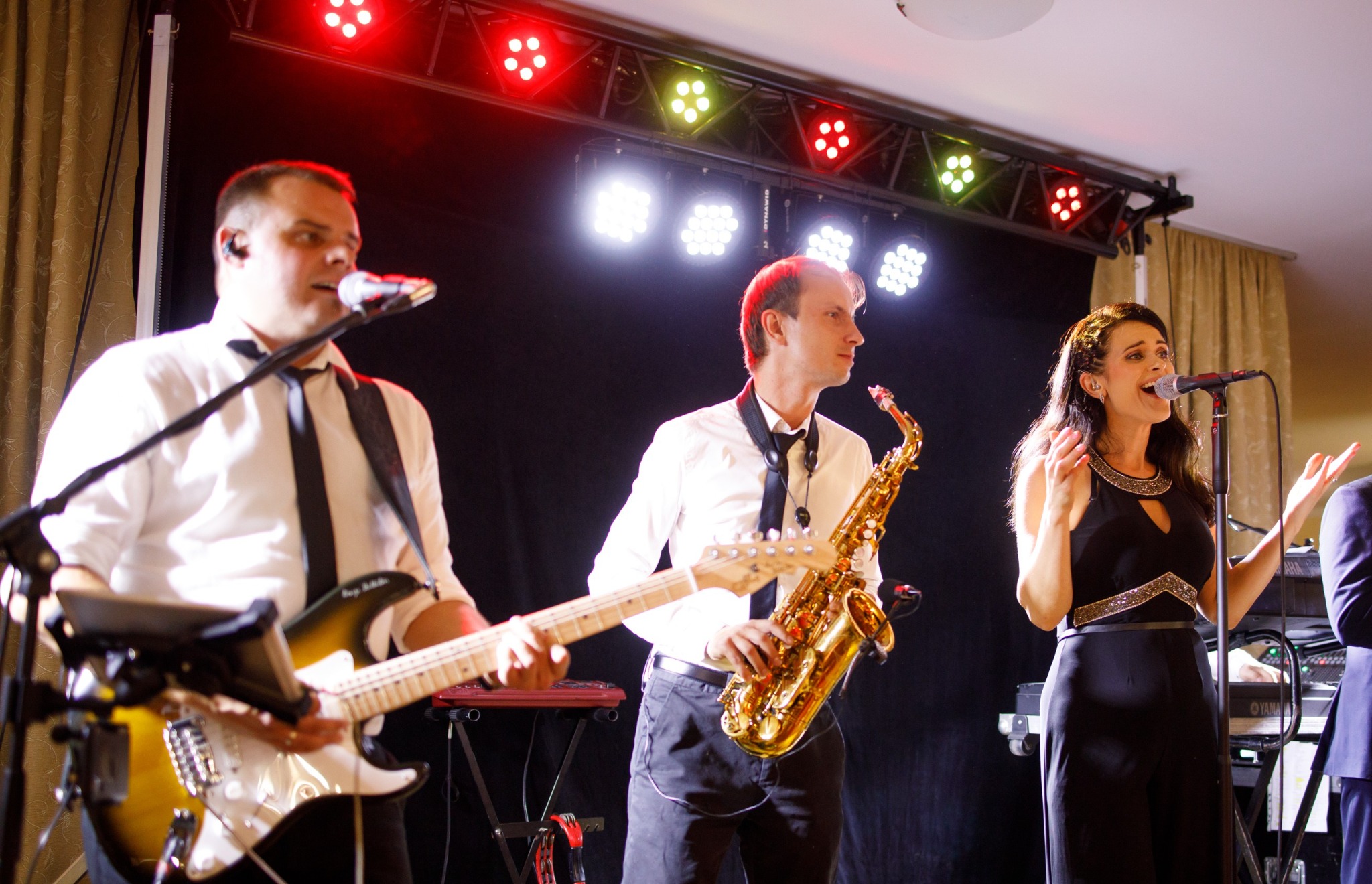 Zespół muzyczny na scenie: gitarzysta z gitarą typu sunburst, saksofonista z saksofonem tenorowym i wokalistka w czarnym kombinezonie, oświetleni reflektorami.