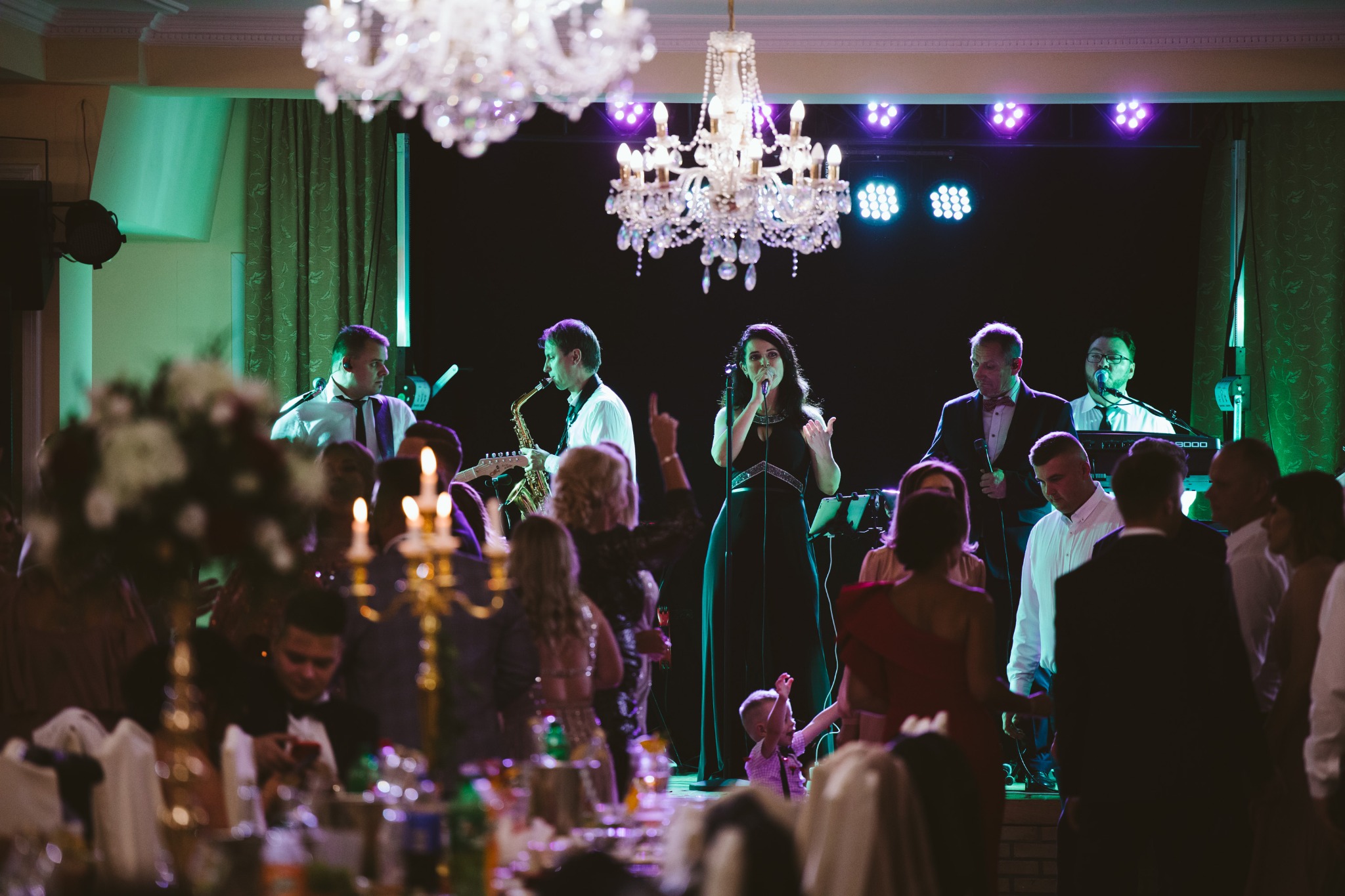 Zespół muzyczny na scenie podczas występu na sali balowej z kryształowymi żyrandolami, oświetleniem scenicznym i publicznością przy stołach.