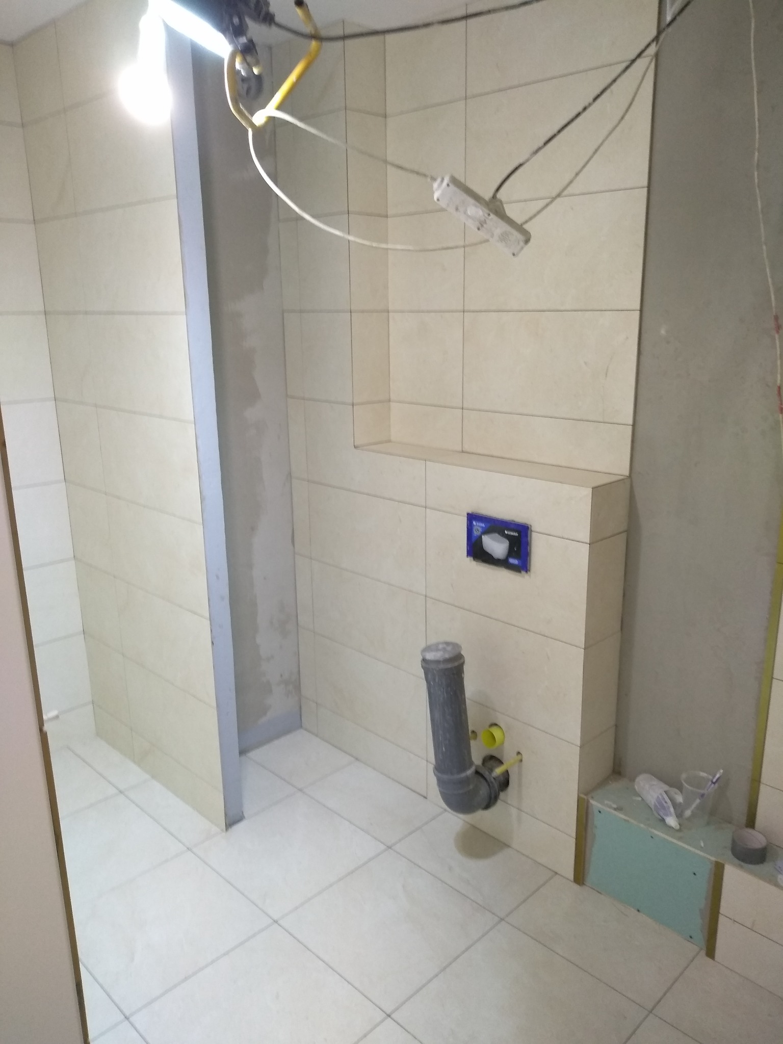 Łazienka w trakcie układania płytek, widoczne rury kanalizacyjne, stelaż podtynkowy WC, narzędzia i materiały budowlane na półce z płyty kartonowo-gipsowej, tymczasowe oświetlenie.