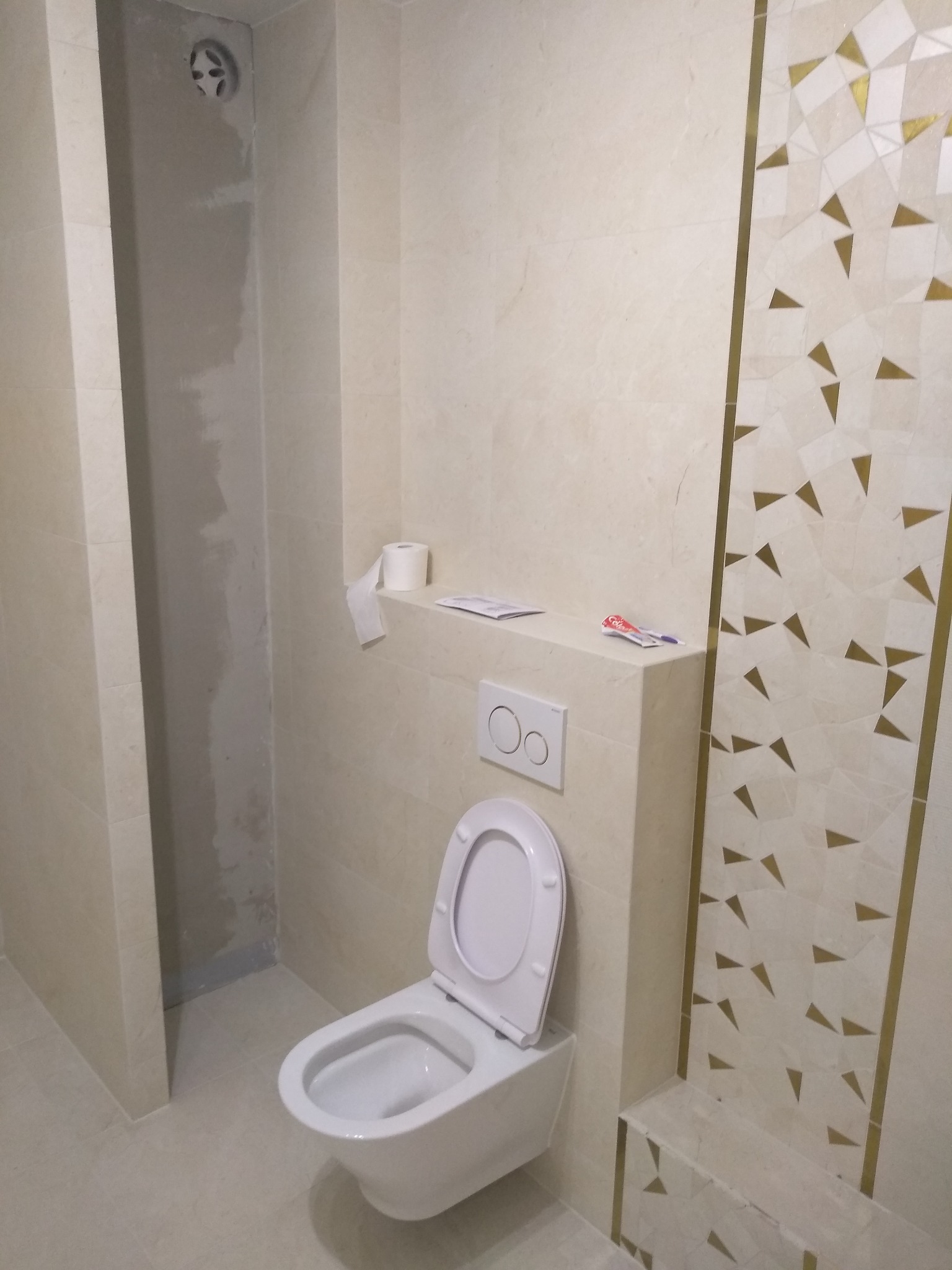 Nowoczesna toaleta podwieszana z otwartą klapą, kremowe płytki z dekoracyjnym motywem geometrycznym w odcieniach bieli i złota, widoczny fragment ściany z niedokończonymi pracami wykończeniowymi...