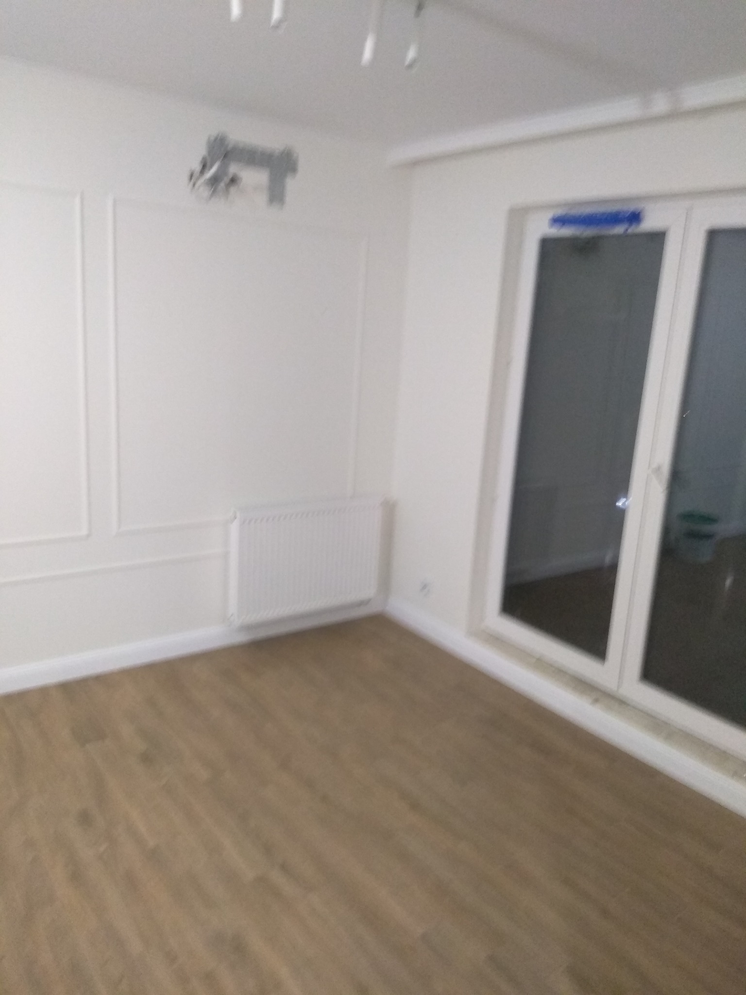 Puste, białe pomieszczenie z panelami podłogowymi, grzejnikiem i dekoracyjnymi listwami ściennymi, przygotowane do dalszych prac.