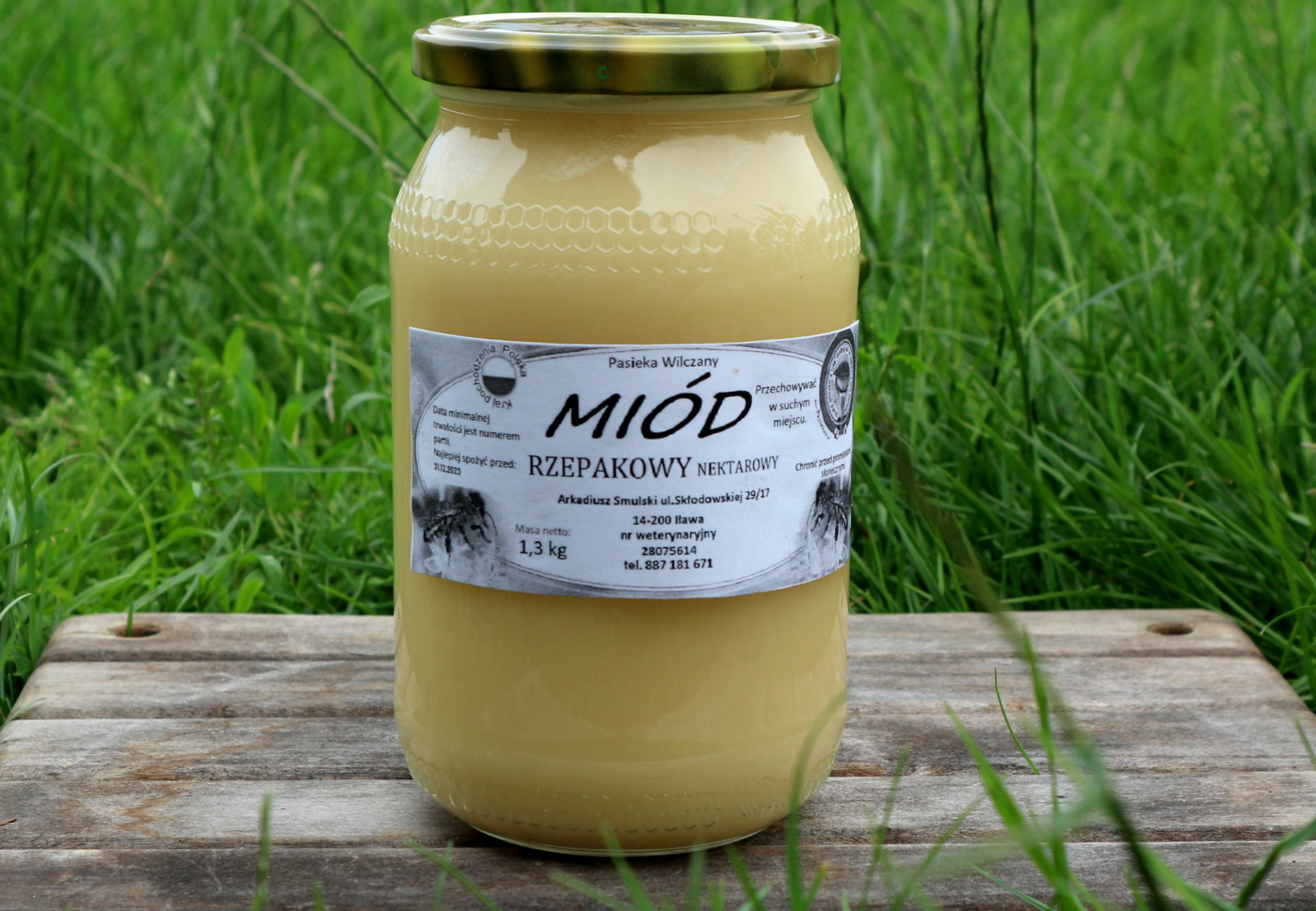 Słoik kremowego, jasnego miodu rzepakowego z etykietą Pasieki Wilczany, umieszczony na drewnianej desce na tle zielonej trawy.