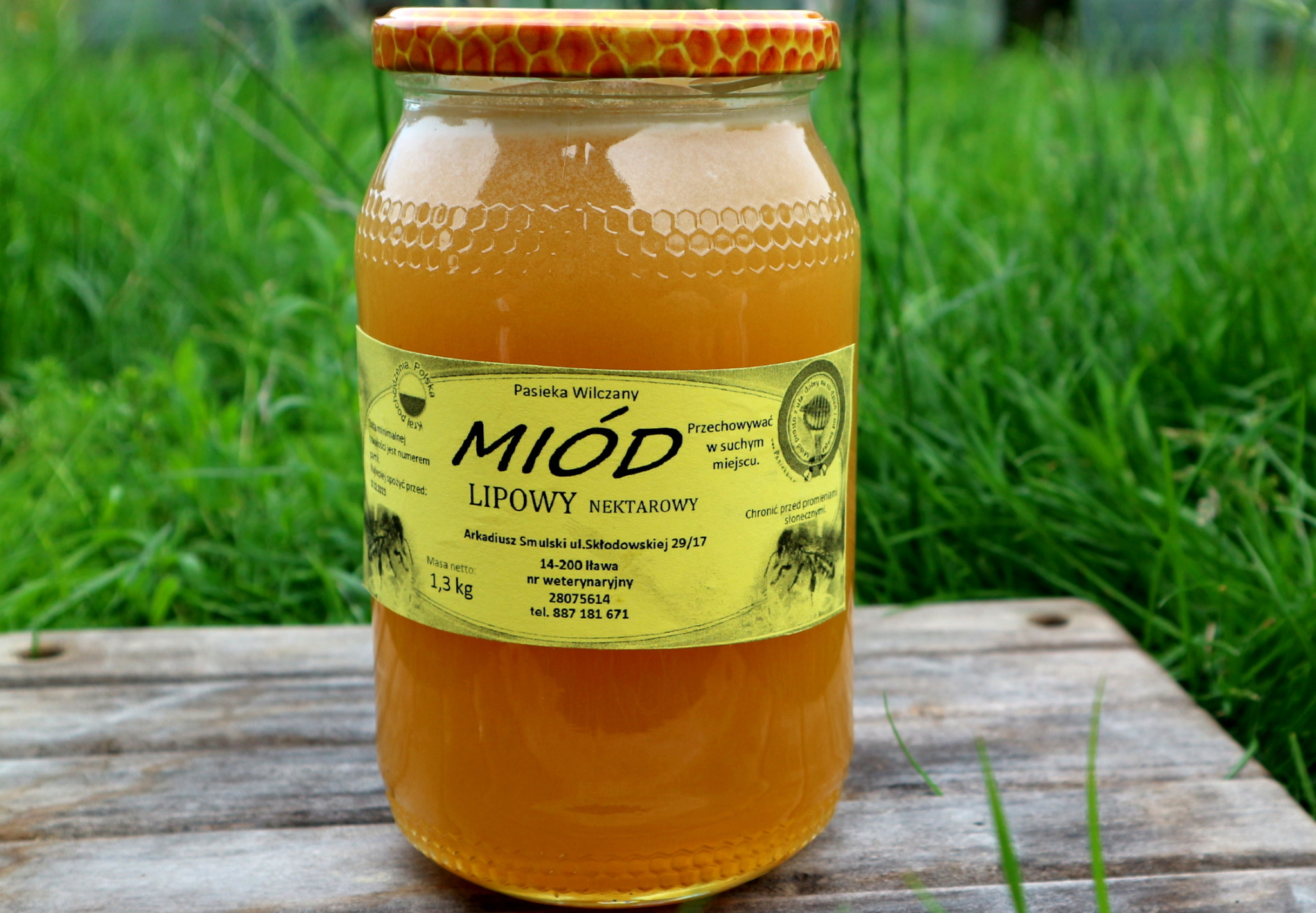 Słoik miodu lipowego nektarowego z etykietą Pasieka Wilczany, ustawiony na drewnianej desce na tle zielonej trawy. Widoczny delikatny wzór plastra miodu na wieczku słoika.