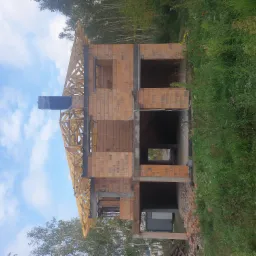 Surowy stan dwukondygnacyjnego domu z cegły ceramicznej z drewnianą konstrukcją dachu, folią na kominie, w tle zieleń, błękitne niebo z chmurami.