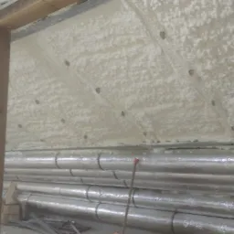 Poddasze po aplikacji izolacji pianką, widoczne rury owinięte folią aluminiową i drewniany element konstrukcyjny.