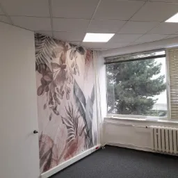 Pokój biurowy z fototapetą w motywy roślinne na ścianie, szara wykładzina, okno z widokiem na drzewo.