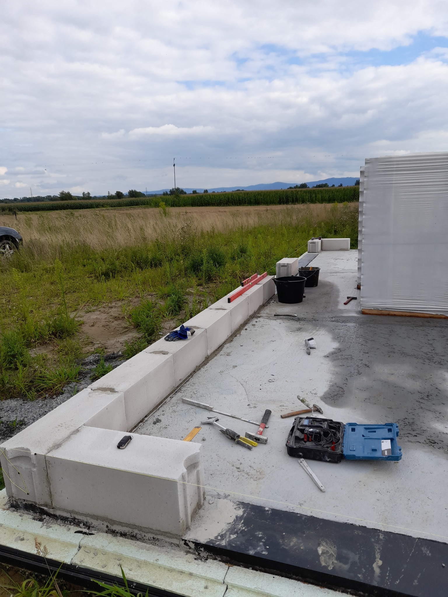 Wznoszenie fundamentów domu z bloczków betonowych, widoczne narzędzia murarskie, poziomica, wiaderka i skrzynka narzędziowa na mokrej betonowej płycie, w tle pole i pochmurne niebo.