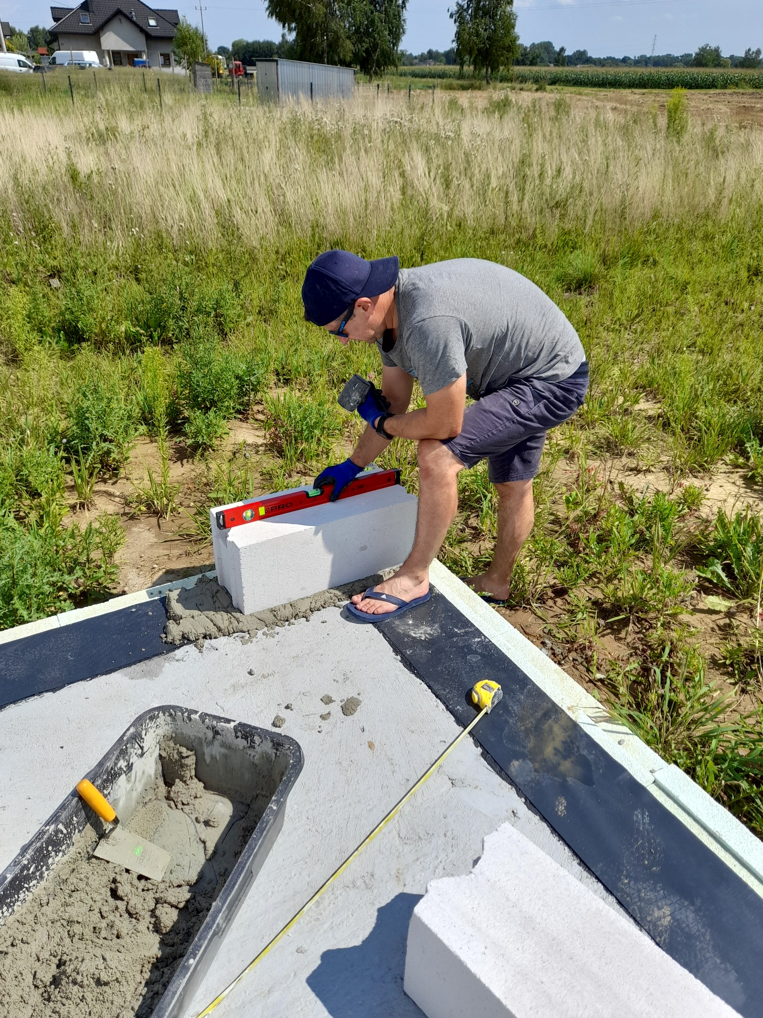 Mężczyzna w kapeluszu i klapkach, w niebieskich rękawicach murarskich, poziomuje bloczek betonowy na fundamencie w otoczeniu wysokiej trawy.