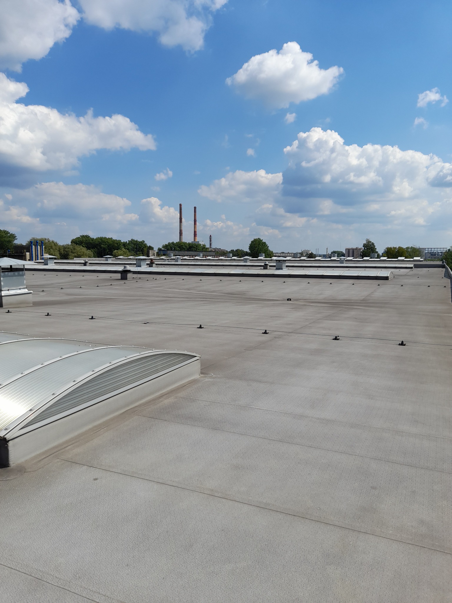 Płaski dach pokryty szarą membraną z widocznymi świetlikami i kominkami wentylacyjnymi, w tle kominy przemysłowe i błękitne niebo z chmurami.