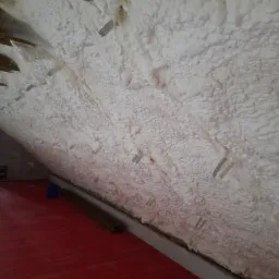 Poddasze z widoczną izolacją natryskową z pianki poliuretanowej, mocowaną metalowymi elementami do krokwi; czerwona, pomalowana podłoga.