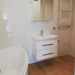 Nowoczesna, jasna łazienka z białą armaturą, umywalką z szafką i grzejnikiem drabinkowym. Ściany wyłożone białymi płytkami z delikatnym wzorem geometrycznym, podłoga z paneli imitujących drewno.