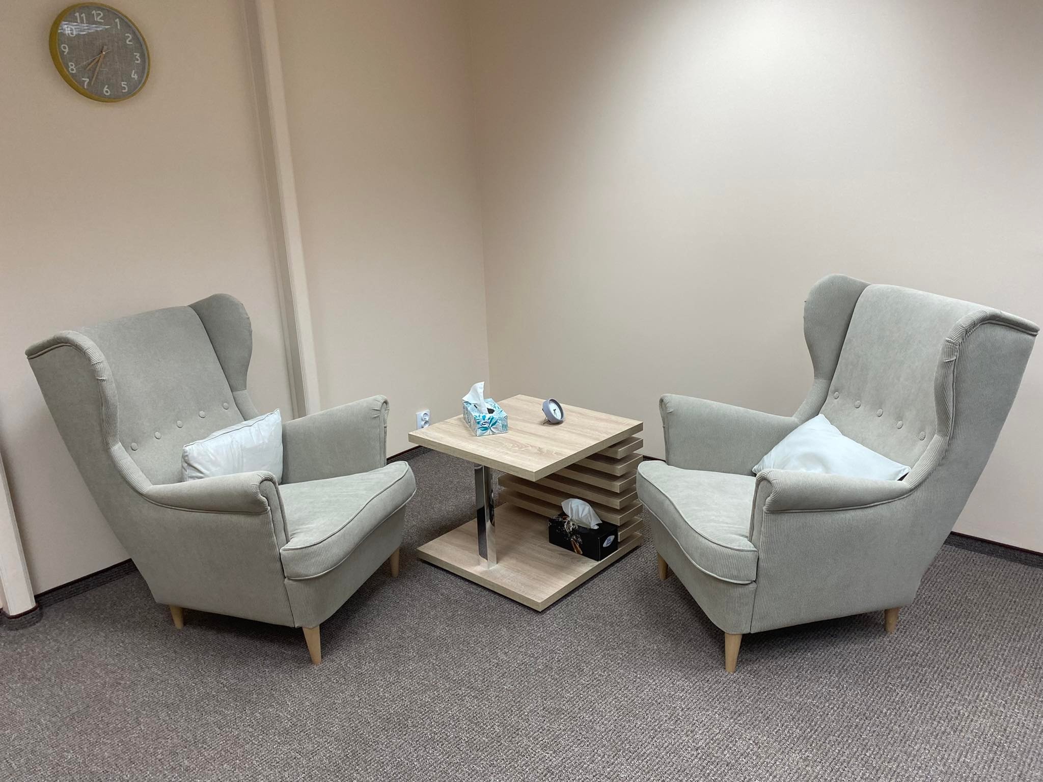 Jasny gabinet z dwoma fotelami uszakami, stolikiem kawowym z chusteczkami i zegarem na ścianie.