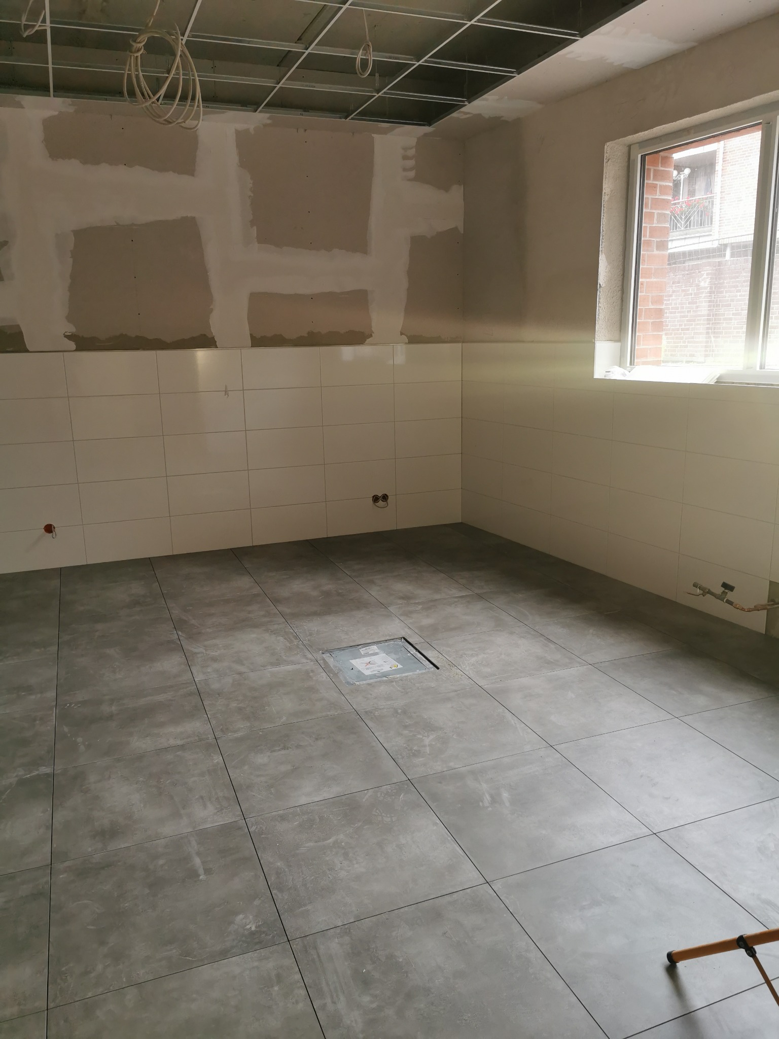 Wnętrze w trakcie remontu: szare płytki podłogowe, białe płytki ścienne do połowy wysokości, surowy sufit podwieszany i ściany przygotowane pod malowanie.