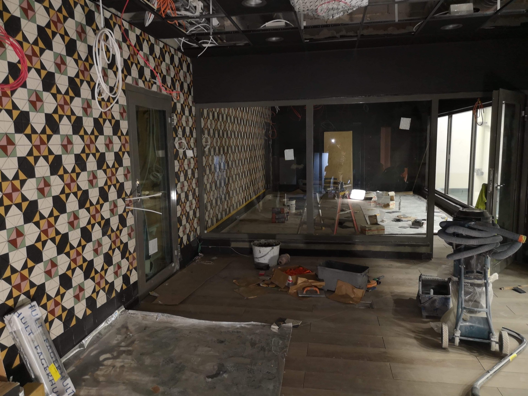 Układanie wzorzystych płytek na ścianie w trakcie remontu lokalu, widoczne narzędzia glazurnicze, kable na suficie i częściowo wykończona podłoga.
